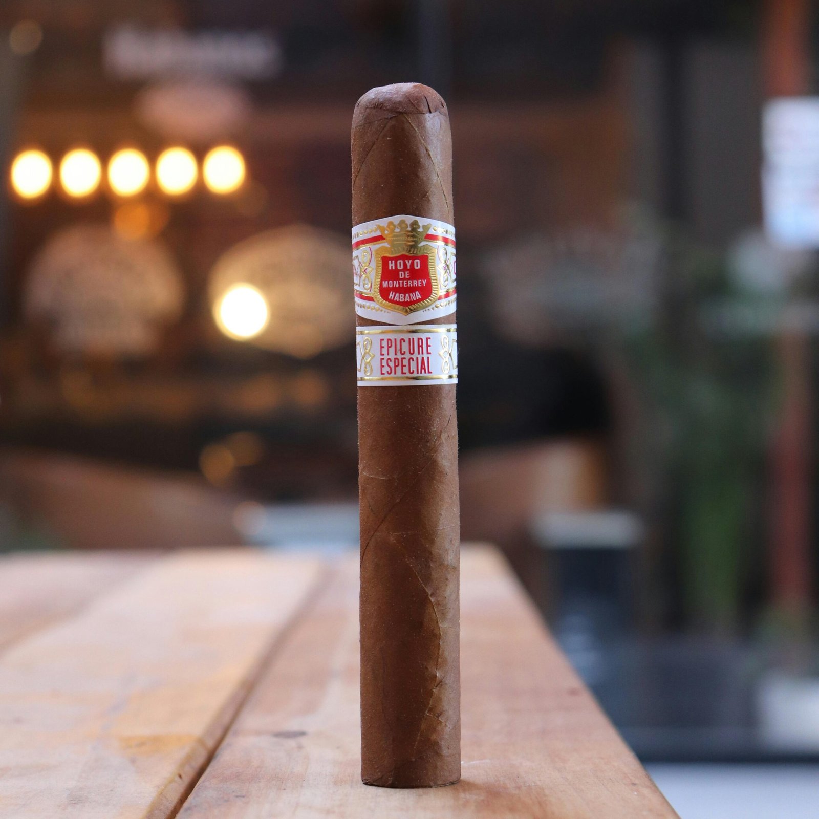 A premium cigar stands on a wooden table in a softly lit indoor setting, creating a warm and luxurious atmosphere.