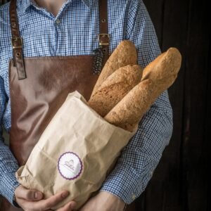 A man in an apron holds fresh baguettes in a paper bag, showcasing artisan baking.